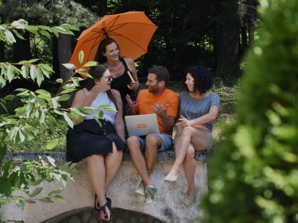 A diverse group of adults enjoying a conversation in nature, sitting with a laptop and umbrella on a sunny day.